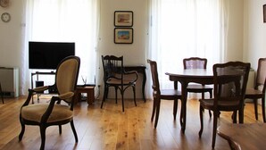 A living room with a television, wooden furniture, and two windows with sheer curtains.