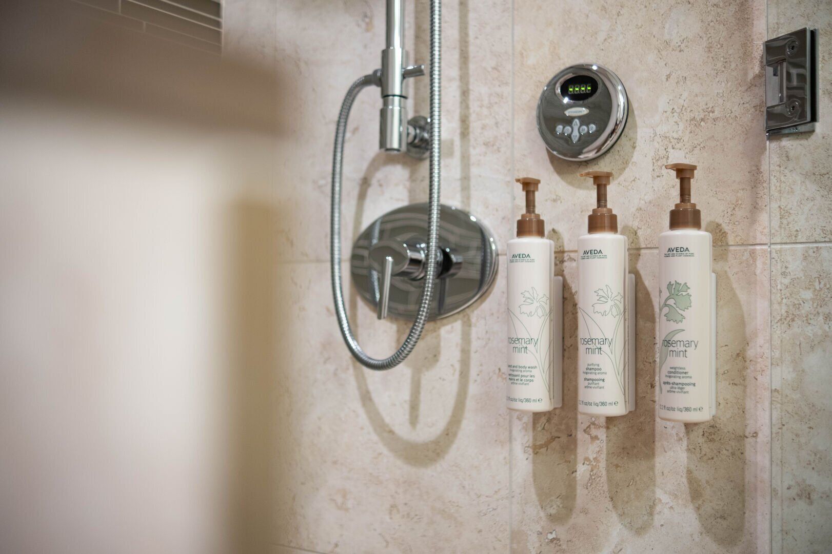 A bathroom with a shower, a digital water temperature control, and three bottles of Aveda Rosemary Mint body wash and conditioner.