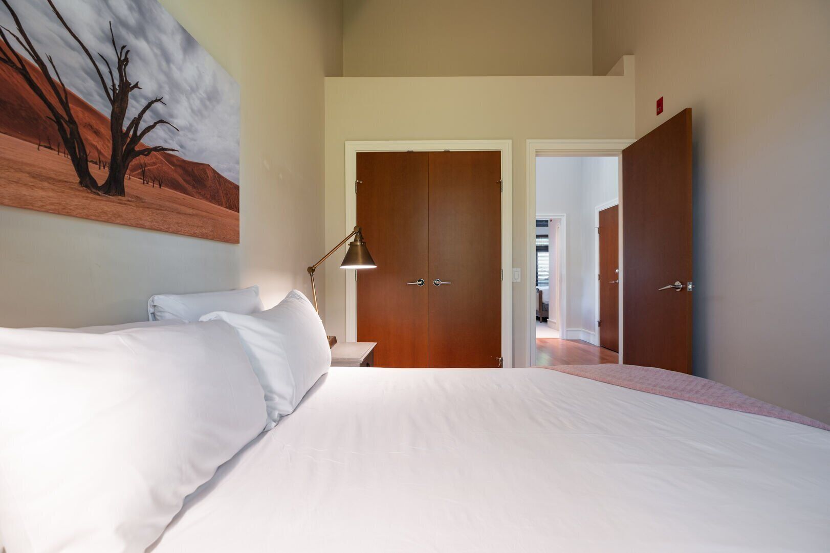 A bedroom with a large bed, a desk with a lamp, and a door leading to another room.