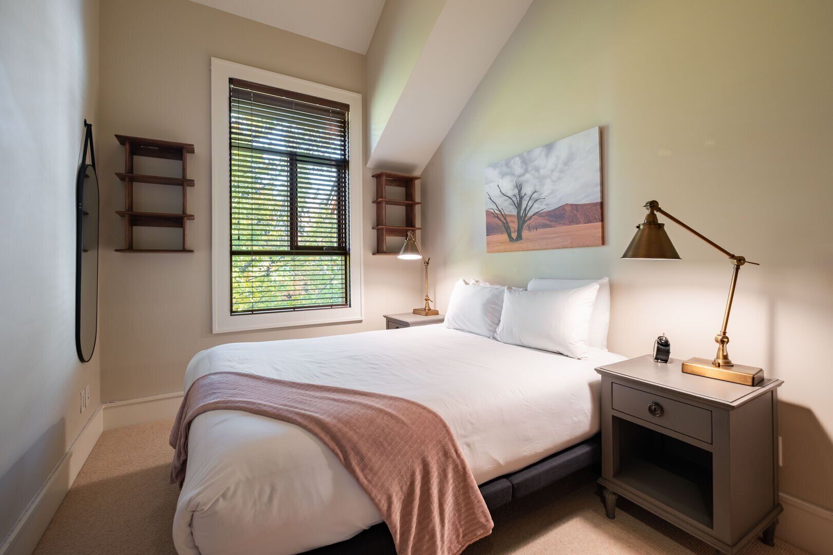 Ein Schlafzimmer mit einem Bett, Nachttischen, einer Lampe, einem Gemälde und einem Fenster mit Jalousien.