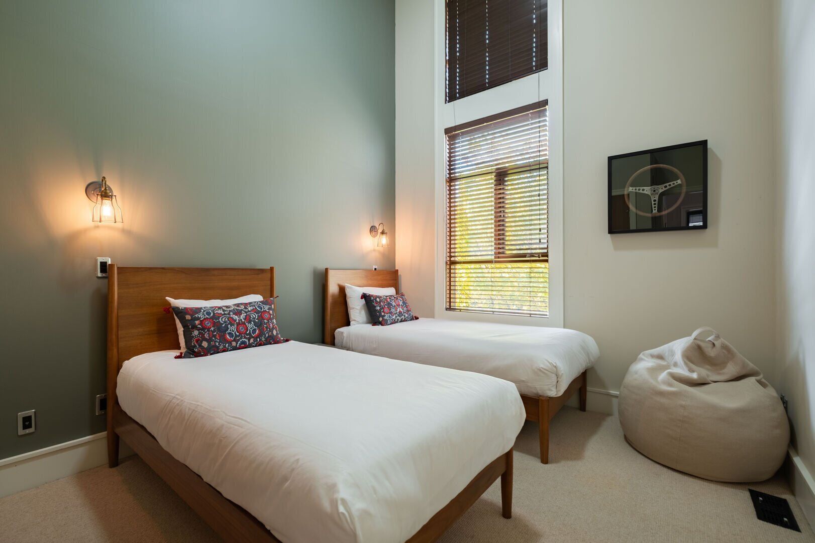 Un dormitorio con dos camas, un sillón de mimbre, una lámpara de pared y una ventana con persianas.