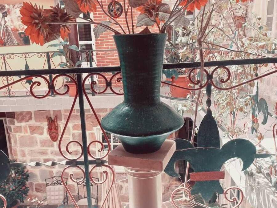 A balcony with a green vase holding red flowers, a metal railing, and a brick wall in the background.