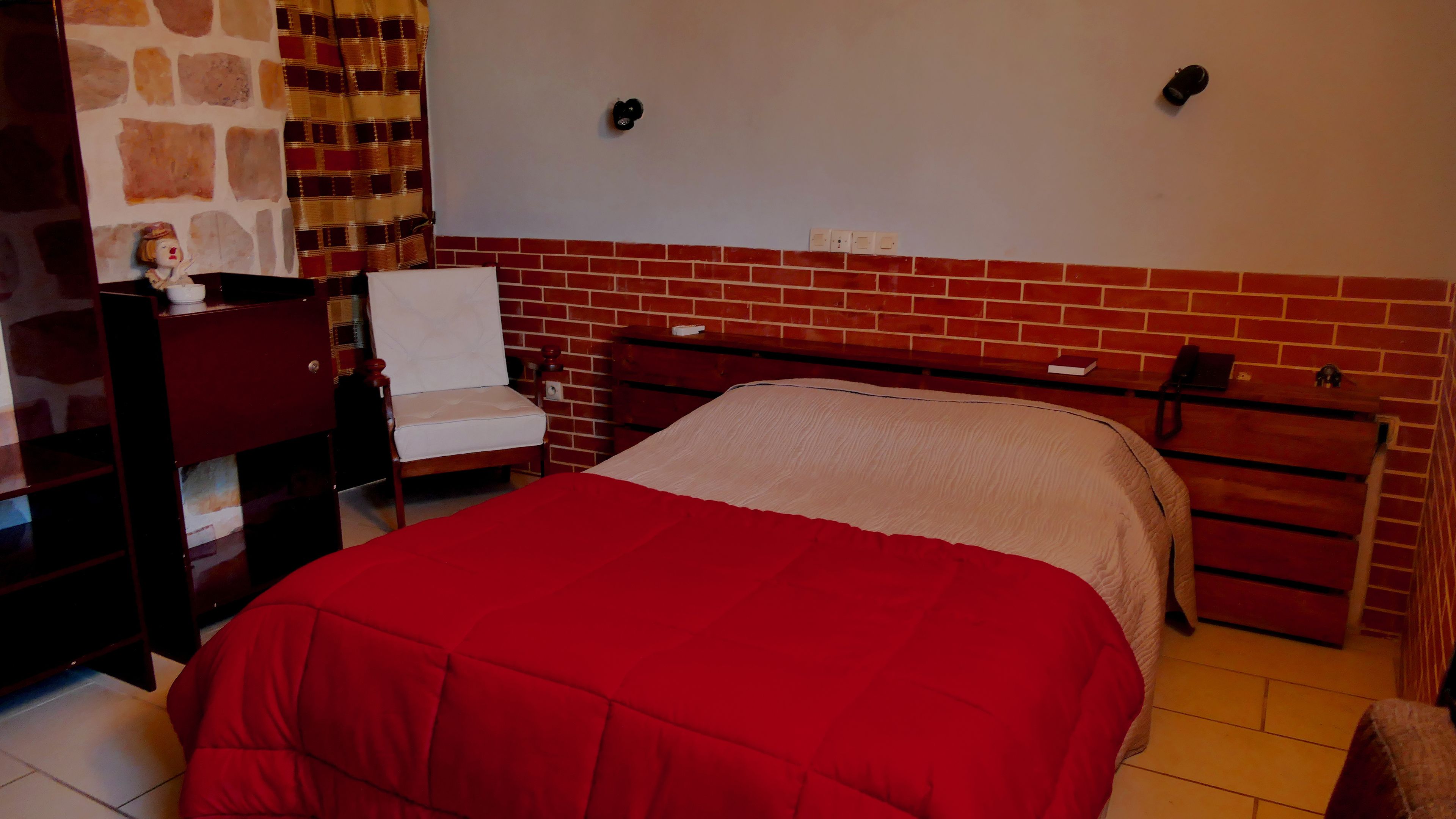 A bedroom with a red bedspread, a wooden nightstand, a chair, a brick-patterned wall, and a stone-patterned wall.