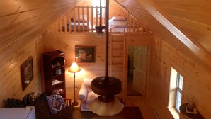A cozy attic bedroom with a sloped ceiling, a wooden ladder, a bed, a chair, a lamp, and a bookshelf.