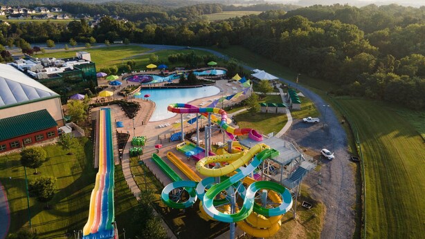 An aerial view of a water park with multiple slides and pools, surrounded by green fields and a distant mountain range.