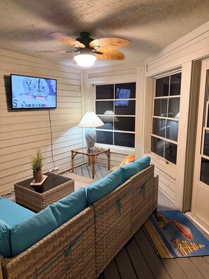 A room with a ceiling fan, a television mounted on the wall, wicker furniture with blue cushions, a small table with a lamp, and a window with multiple panes. - A+Coastal Escape: Beach, Golf, Tennis Club Access Available (Seabrook Island)