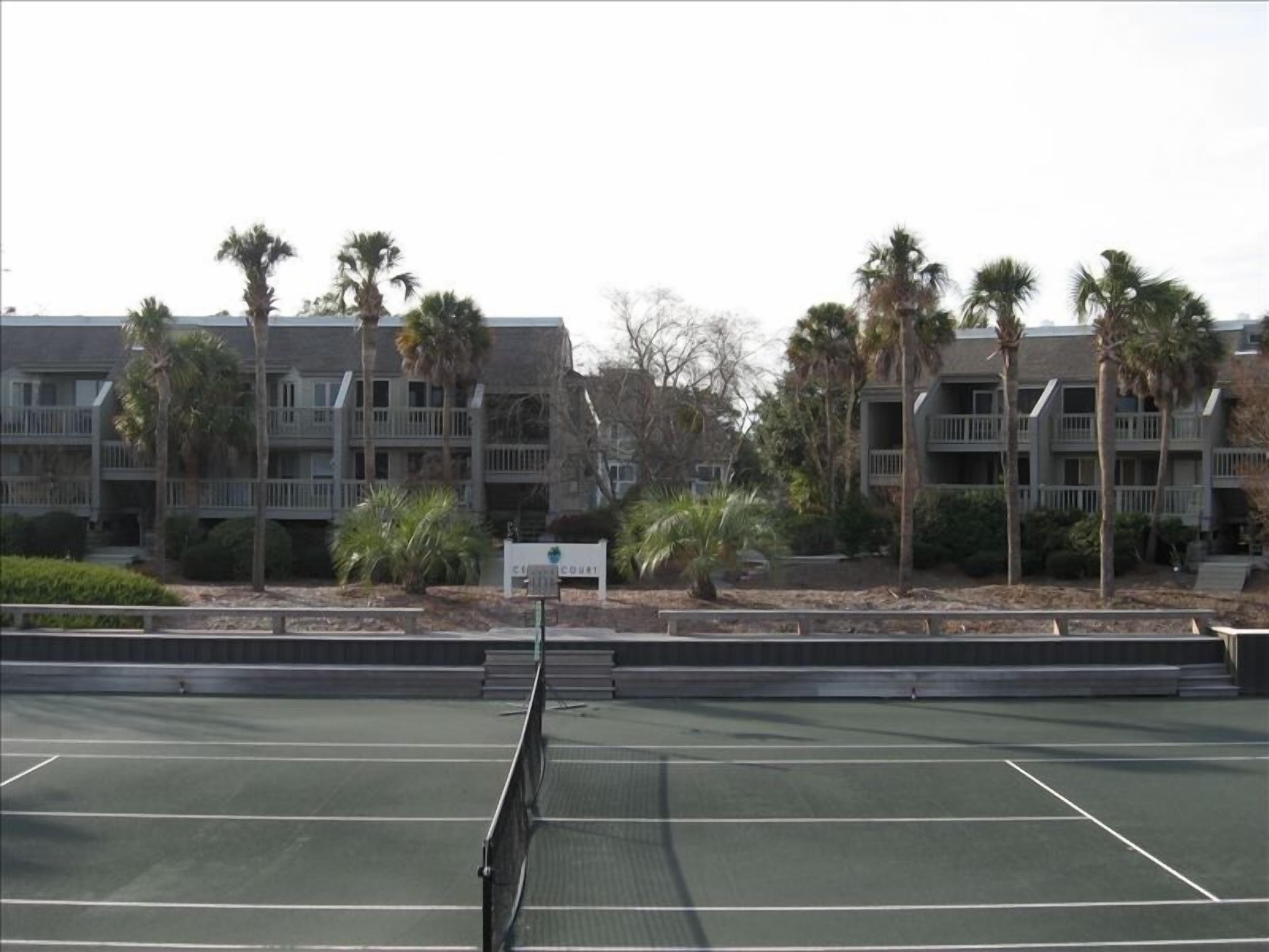 Una cancha de tenis con una red en el centro, rodeada por un edificio con balcones y palmeras.