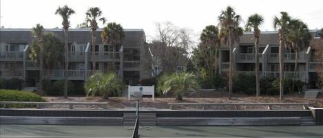 Una cancha de tenis con una red en el centro, rodeada por un edificio con balcones y palmeras.