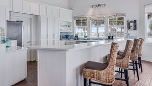 A modern kitchen with white cabinetry, a large island with bar stools, and a view of palm trees through the windows. - Sunsets | Private Pool & Hot Tub | Green Lights | Boat Lift | Icemaker (Rockport)
