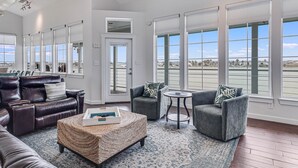 A spacious living room with a leather sofa, a grey armchair, and a round coffee table. Large windows offer a view of the outdoors. - Sunsets | Private Pool & Hot Tub | Green Lights | Boat Lift | Icemaker (Rockport)