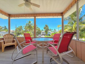 A covered outdoor seating area with a table and chairs, large windows with a view of palm trees and houses, and a ceiling fan. - 231 Dundee Cozy Home Short Walk to Beach (Fort Myers Beach)