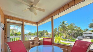 A room with a glass table and red chairs, a ceiling fan, and a view of a residential street with palm trees. - 231 Dundee Cozy Home Short Walk to Beach (Fort Myers Beach)