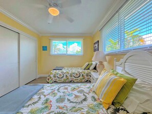 Une chambre avec deux lits, un ventilateur de plafond, une fenêtre avec des stores et une vue sur la verdure.