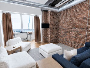 A modern living room with a brick wall, a flat-screen TV mounted on the wall, and a balcony with a view of the city.