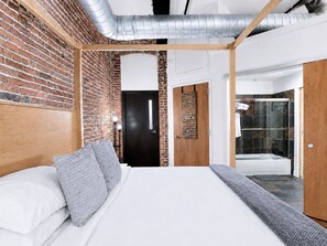 Quarto com parede de tijolos, cabeceira de madeira, cama com roupa de cama branca e área de chuveiro com porta de vidro.