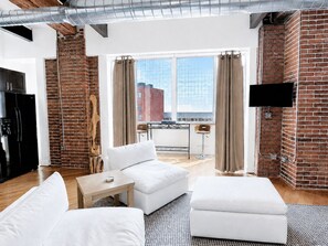 Uma sala de estar moderna com parede de tijolos, uma geladeira preta, sofá branco, mesa de centro de madeira e uma janela grande com cortinas.