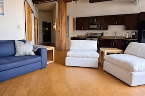 Uma sala de estar com um sofá azul, dois pufes brancos e uma mesa de centro de madeira. Uma cozinha é visível ao fundo, com armários de madeira escura e eletrodomésticos modernos.