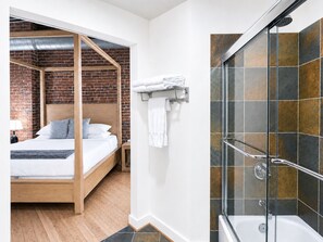 A modern bedroom with a wooden four-poster bed, a brick wall, and a glass-enclosed shower area.