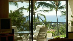 Habitación con un amplio ventanal corredizo que da a un balcón con vistas a palmeras y al océano. Hay una mesa con sillas y un televisor sobre un mueble.