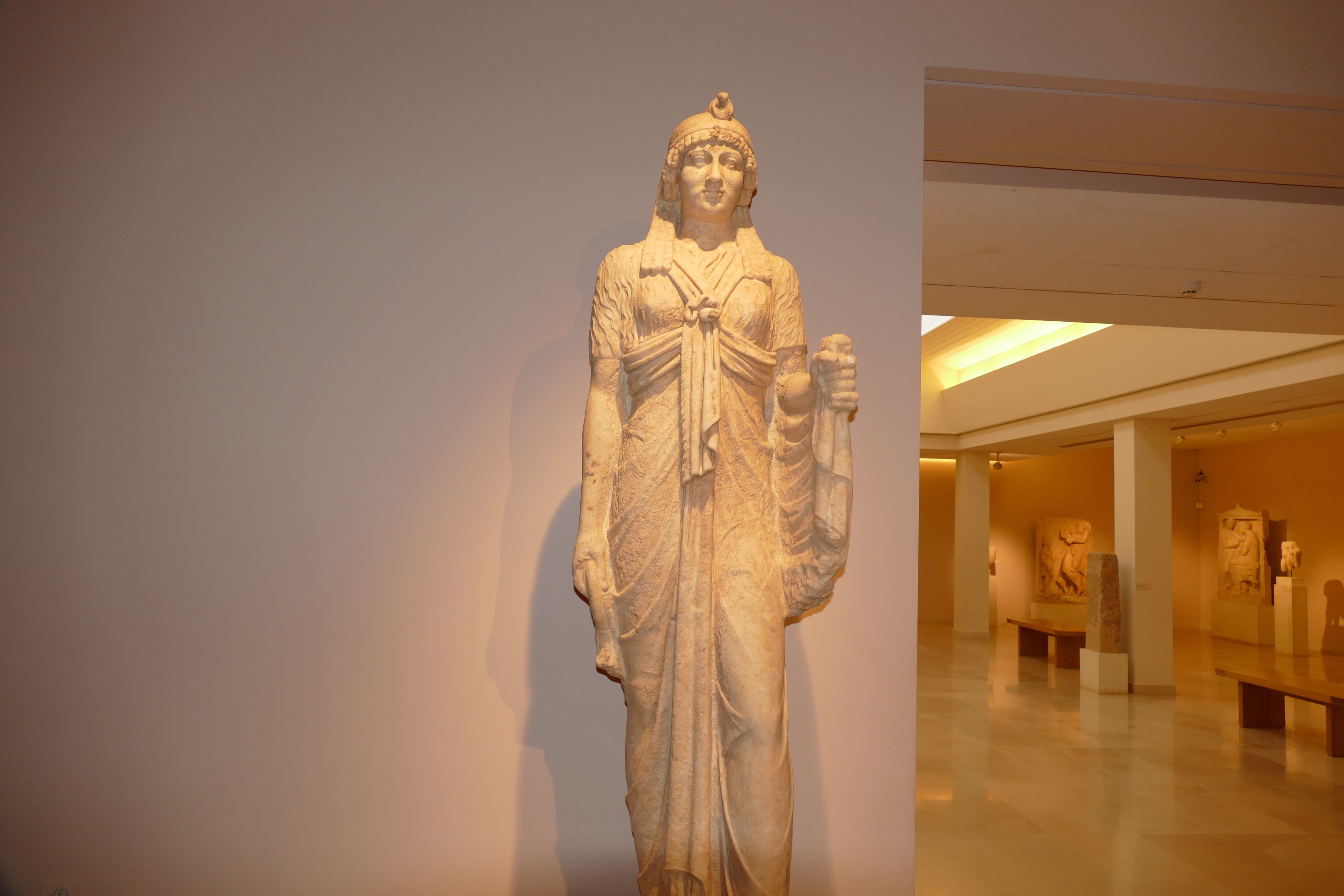 Eine Marmorstatue in einem Museum, vor einem schlichten Hintergrund und mit weiteren Statuen im Hintergrund.