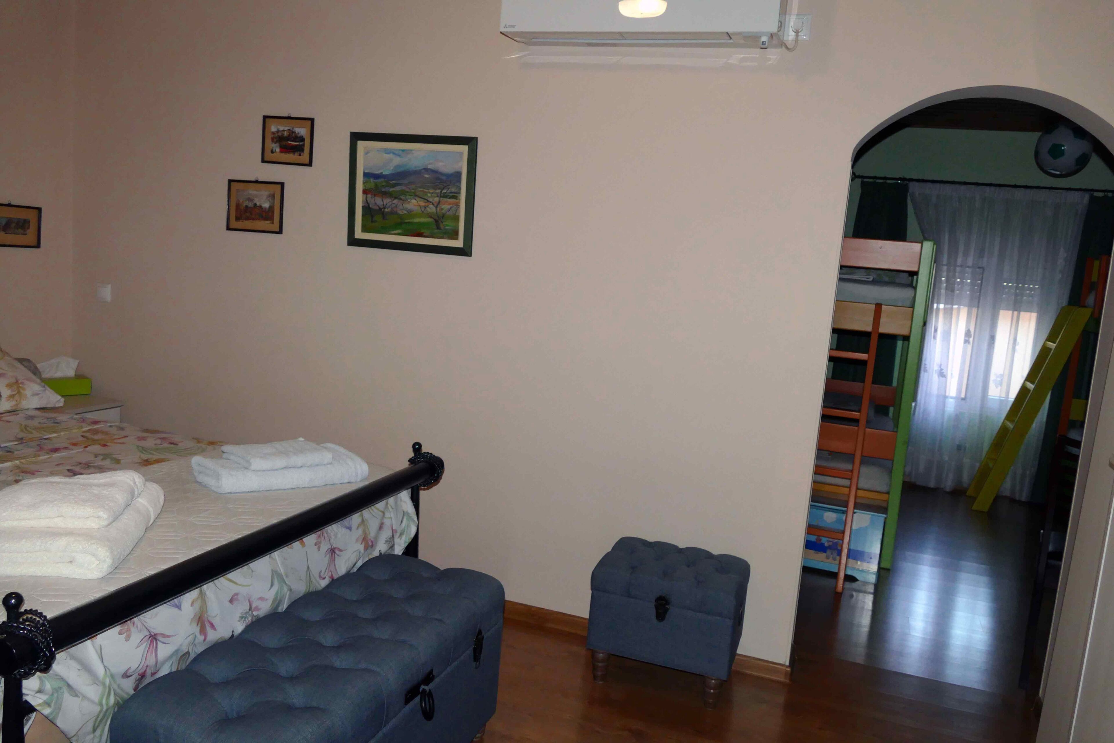 Un dormitorio con una cama, un taburete pequeño y una cama litera al fondo.