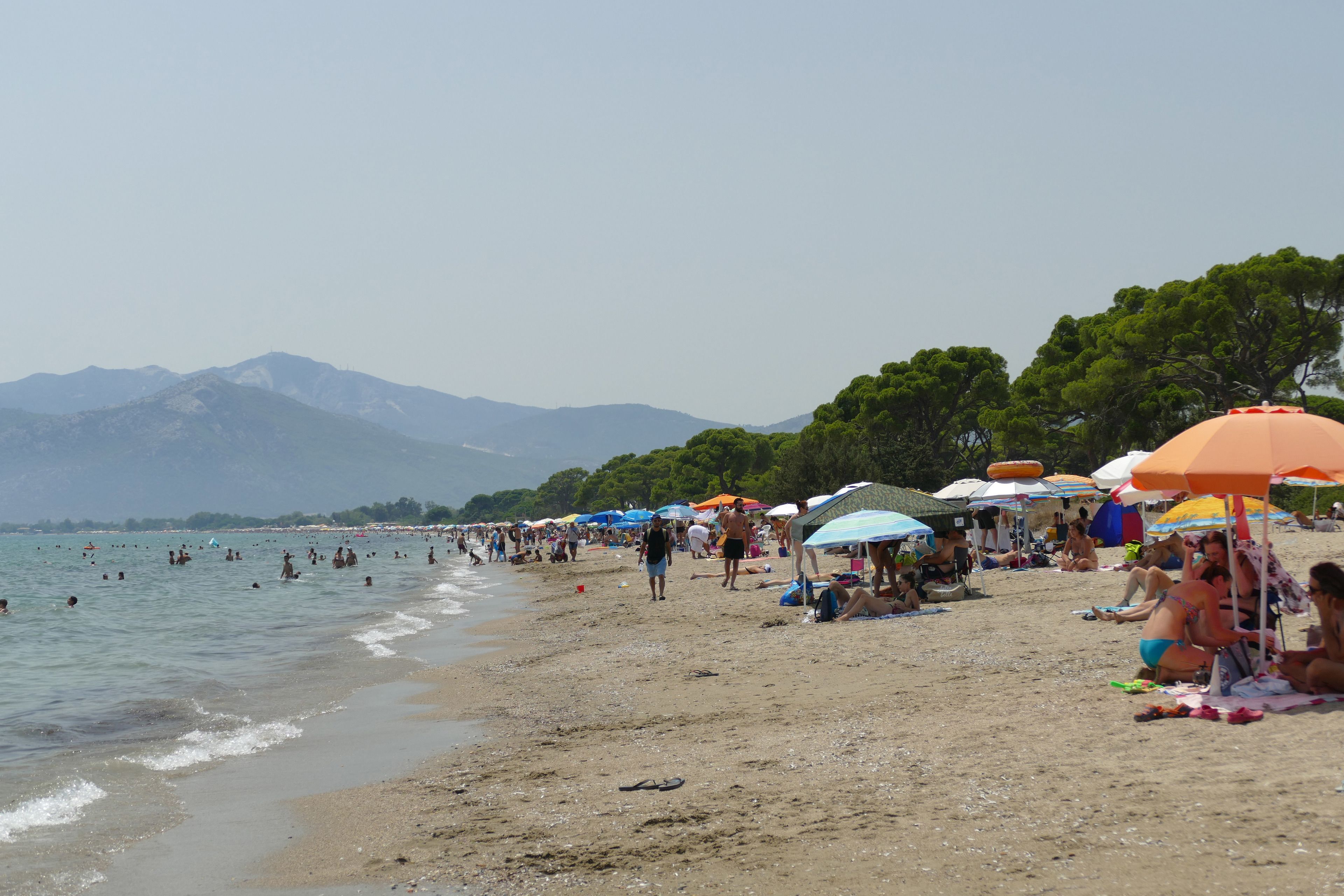 A beach with people sunbathing and swimming, umbrellas, and mountains in the background.
