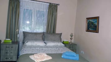 A neatly made bed with a floral bedspread, two pillows, and a blue towel. There is a nightstand with a lamp and a green box. The room has a window with curtains and a painting on the wall.