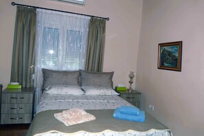 A neatly made bed with a floral bedspread, two pillows, and a blue towel. There is a nightstand with a lamp and a green box. The room has a window with curtains and a painting on the wall.