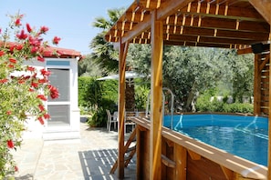 A wooden pool deck with a pool, surrounded by lush greenery and a white building with a red roof.
