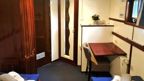 Una stanza piccola con un letto, un tavolino e una sedia. Si nota un termosifone e uno specchio a parete.