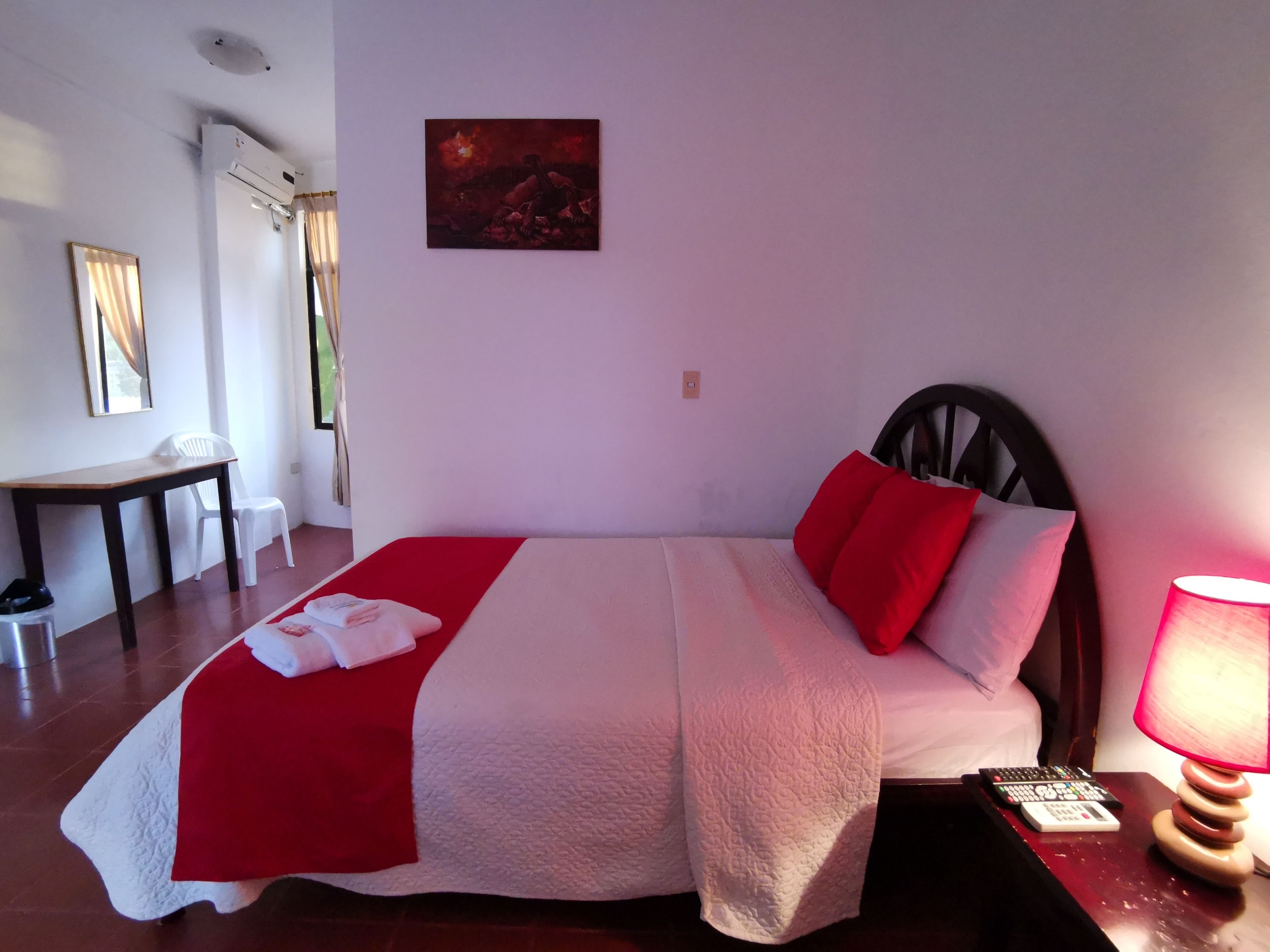 A neatly made bed with a red and white bedspread, a wooden headboard, and a red pillow. There is a small table with a chair next to the bed, a lamp on the nightstand, and a painting on the wall.