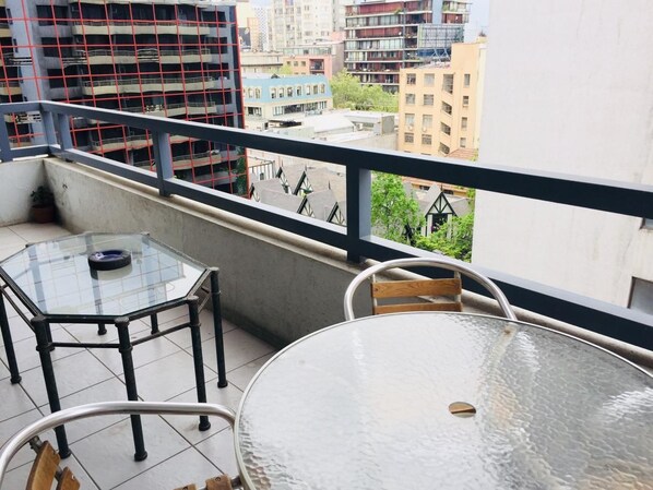 Varanda com mesa e cadeiras de vidro, guarda-corpo de metal e vista para edifícios ao redor.