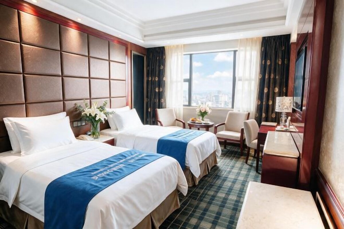 A hotel room with two beds, a sitting area with a chair and table, and a large window with a city view.