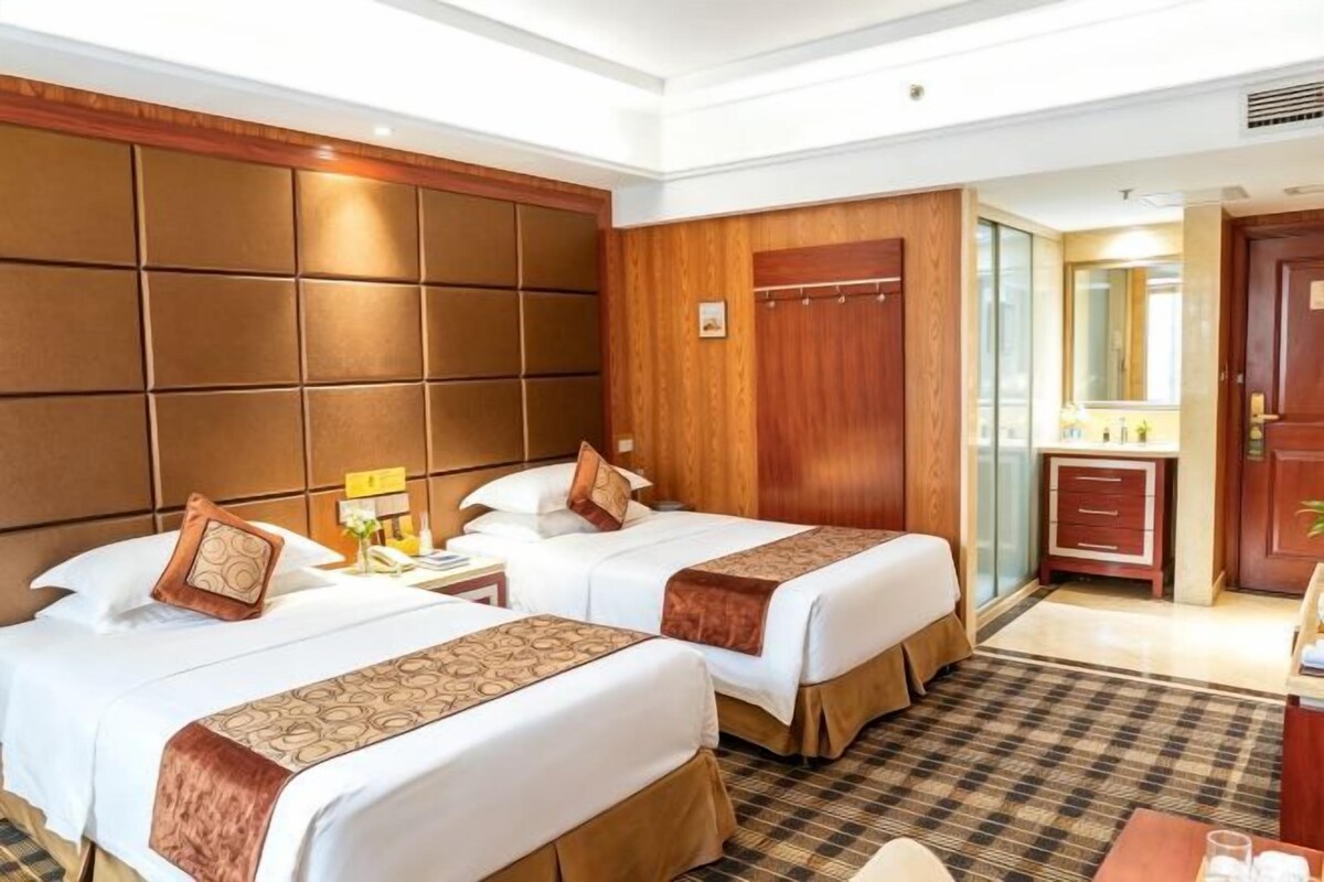 A hotel room with two beds, a wooden headboard, a small bedside table, a mirror, and a bathroom with a sink and mirror.
