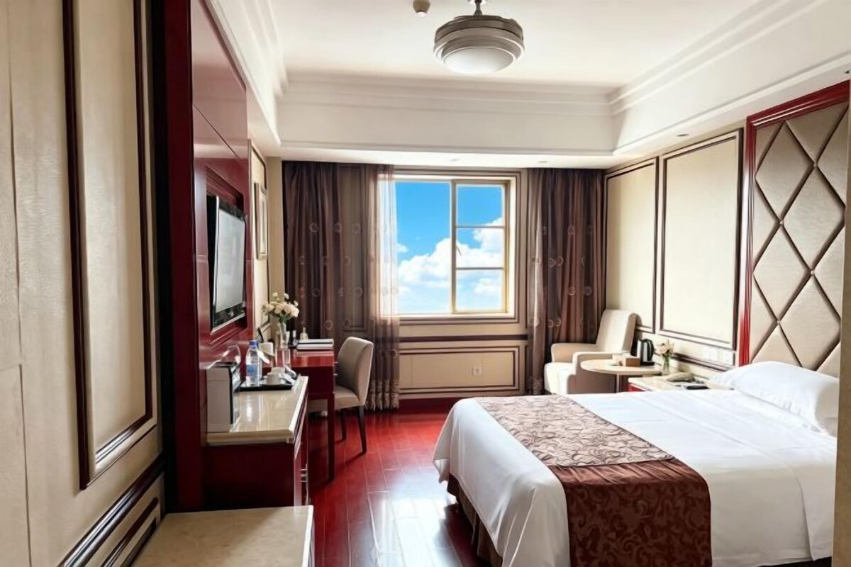 A hotel room with a bed, a desk, a chair, a small table, a mirror, and a window with curtains.