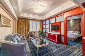 A hotel room with a sofa, armchair, TV, and a bed with a red cabinet.