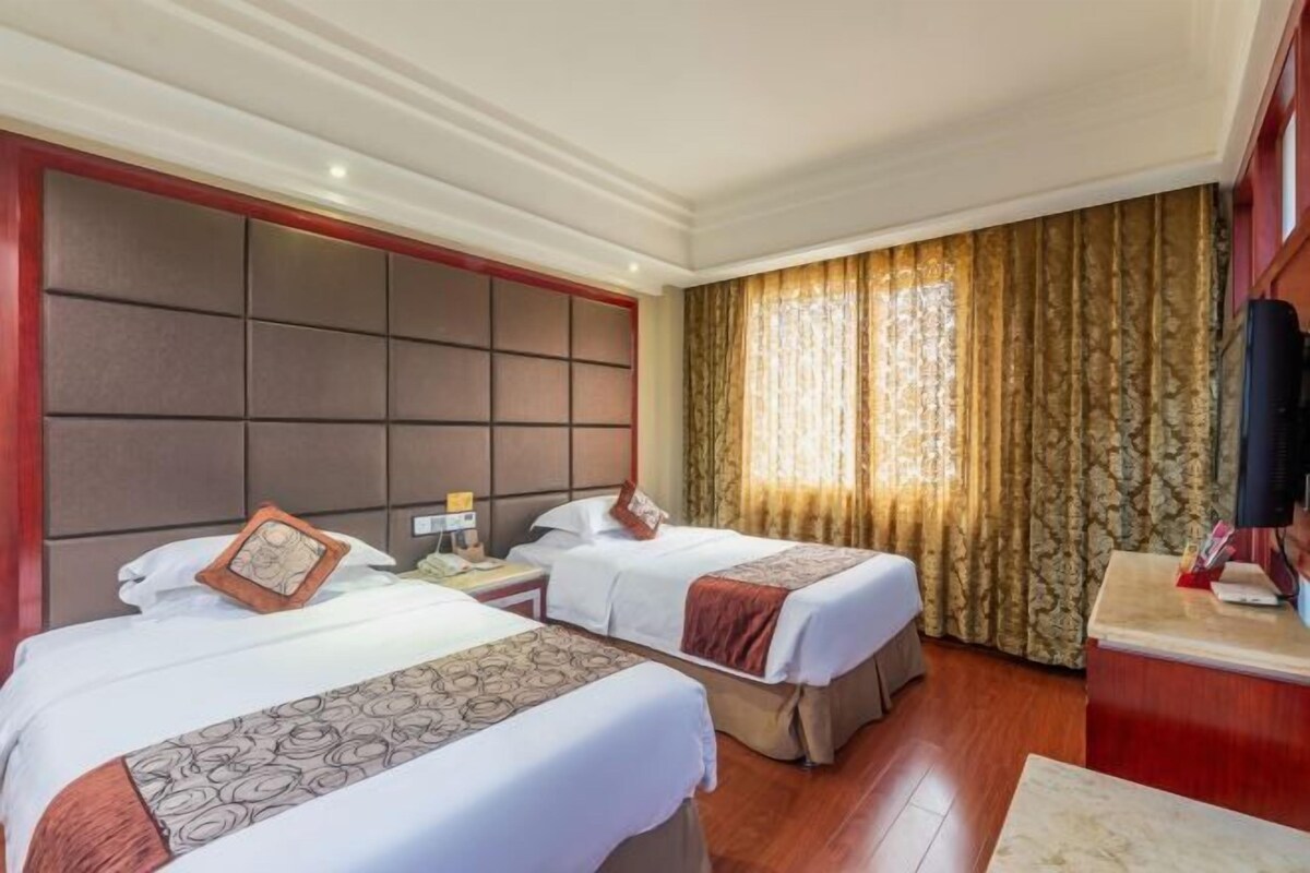 A hotel room with two beds, a large padded headboard, a wooden desk, and a window with curtains.