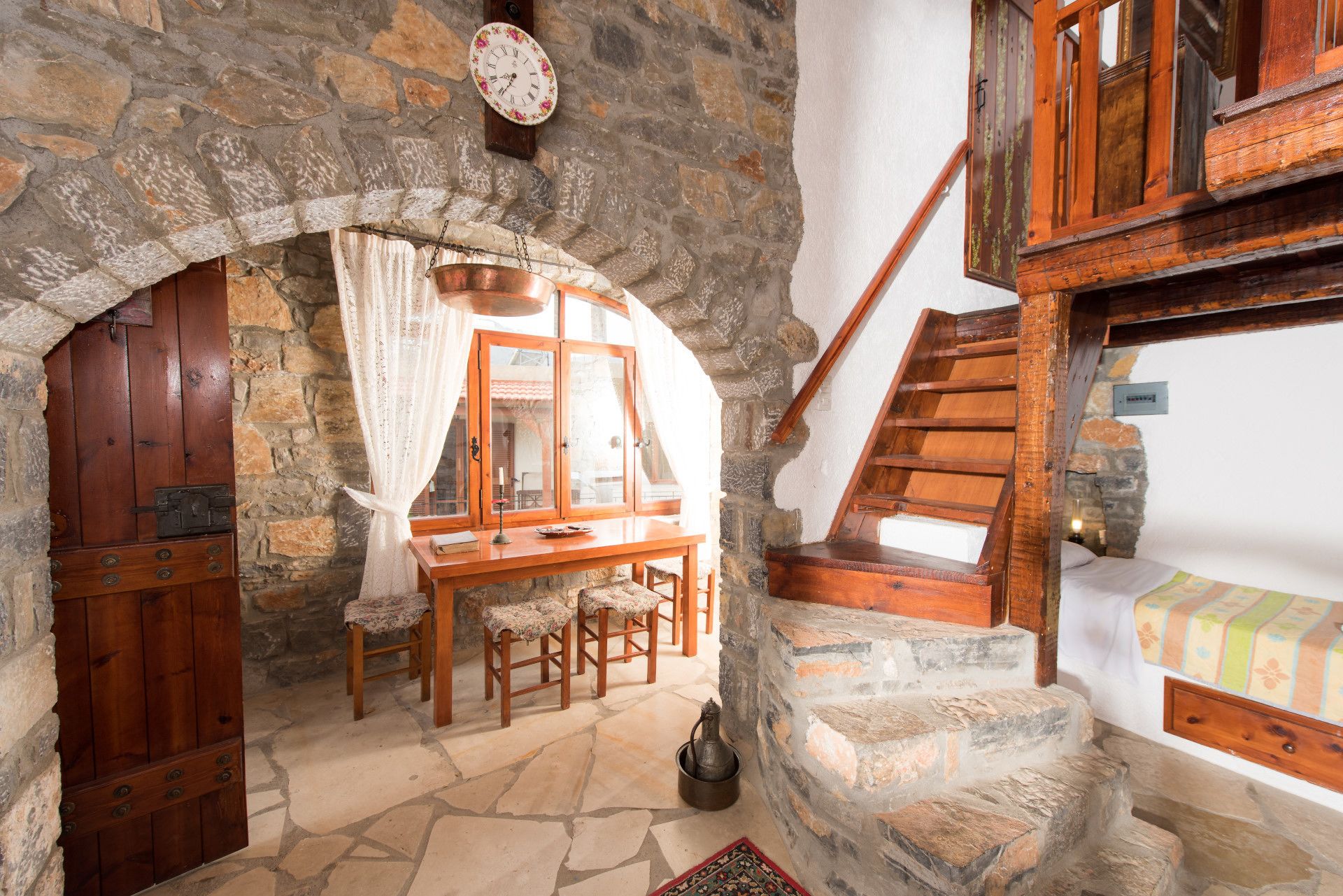Una habitación rústica con paredes de piedra, una mesa de madera con sillas, una escalera de madera y una cama con una manta colorida.
