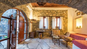 A rustic stone-walled room with a wooden door, a stone floor, a table with a chair, and a bed with a red blanket.