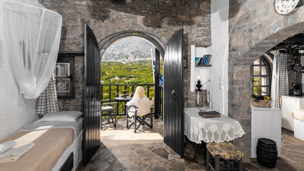 A rustic room with a bed, a table, and a view of a mountainous landscape through an open archway.