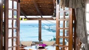 A view from a wooden-framed window overlooking a coastal town with a bridge. Inside, there is a round table with chairs and a vase of flowers.