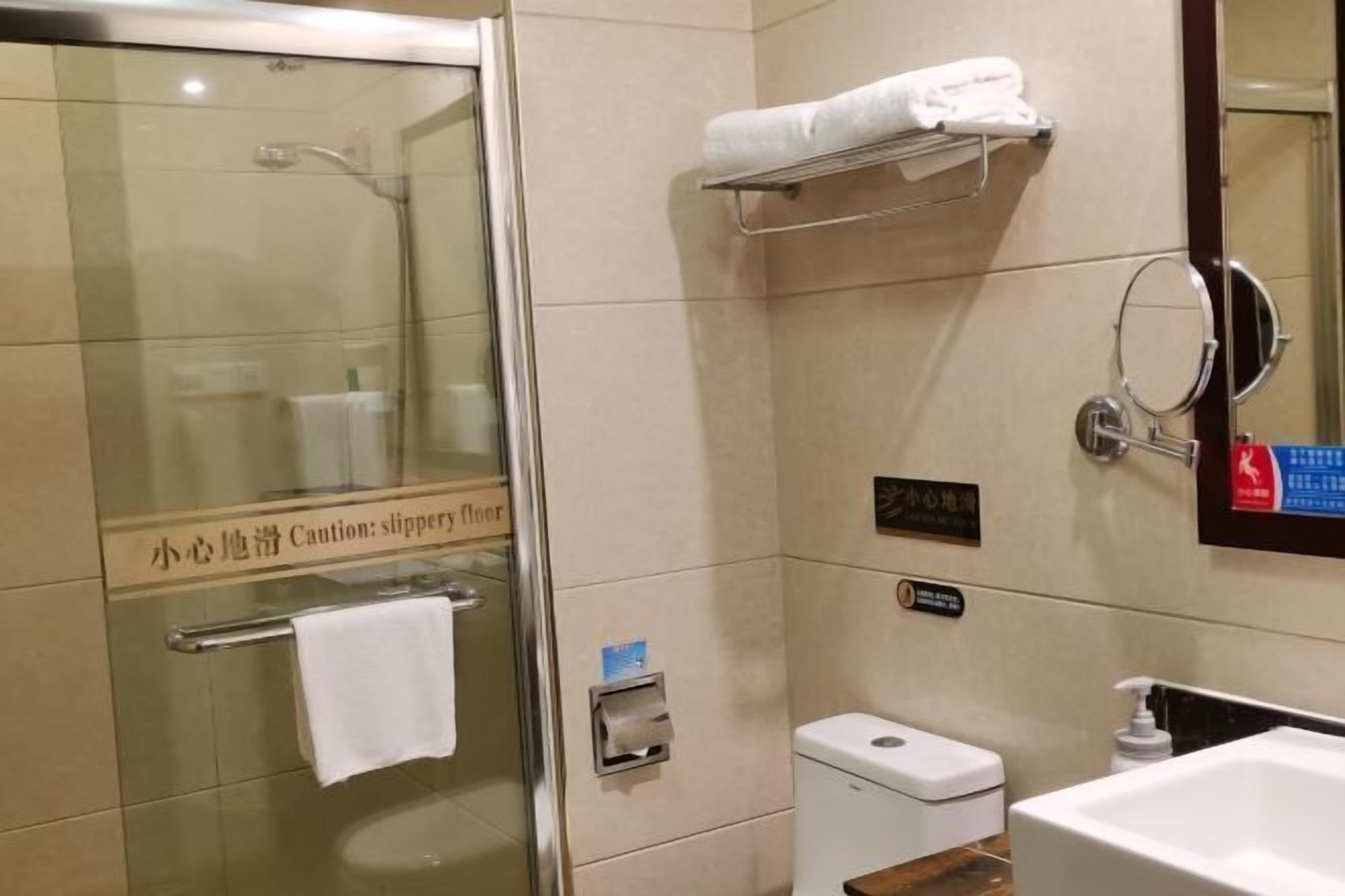 A hotel bathroom with a glass shower enclosure, a toilet, a sink, and a towel rack.