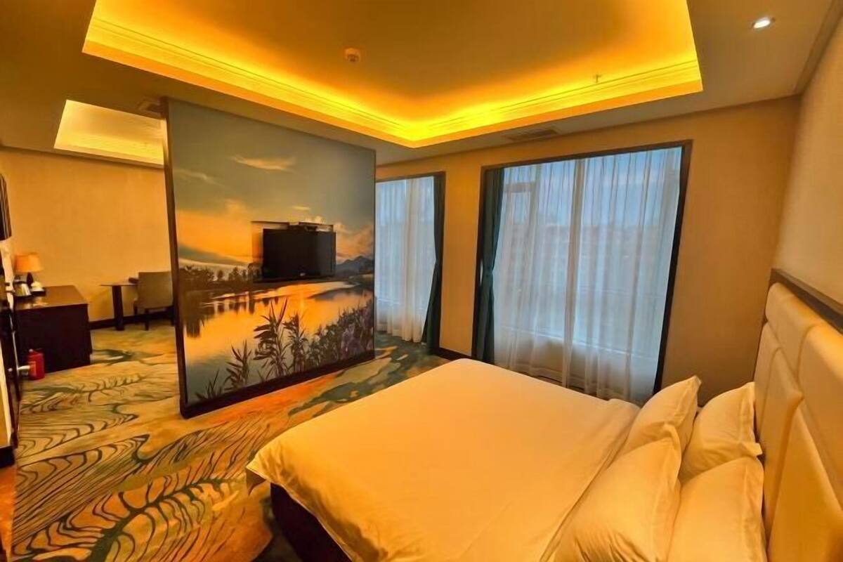 A hotel room with a bed, a painting on the wall, a desk with a lamp, and a view of the outdoors through the window.