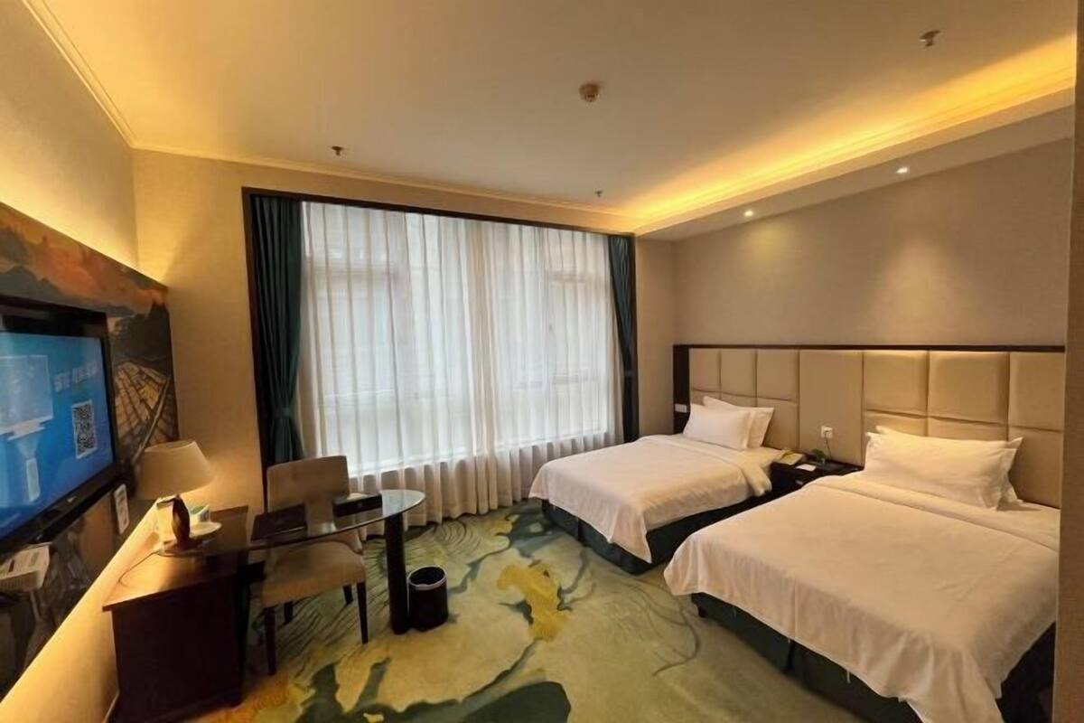 A hotel room with two beds, a desk, a television, and a large window with curtains.
