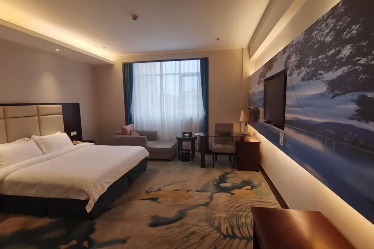 A hotel room with a large bed, a desk, a chair, a small table, and a large window with sheer curtains.