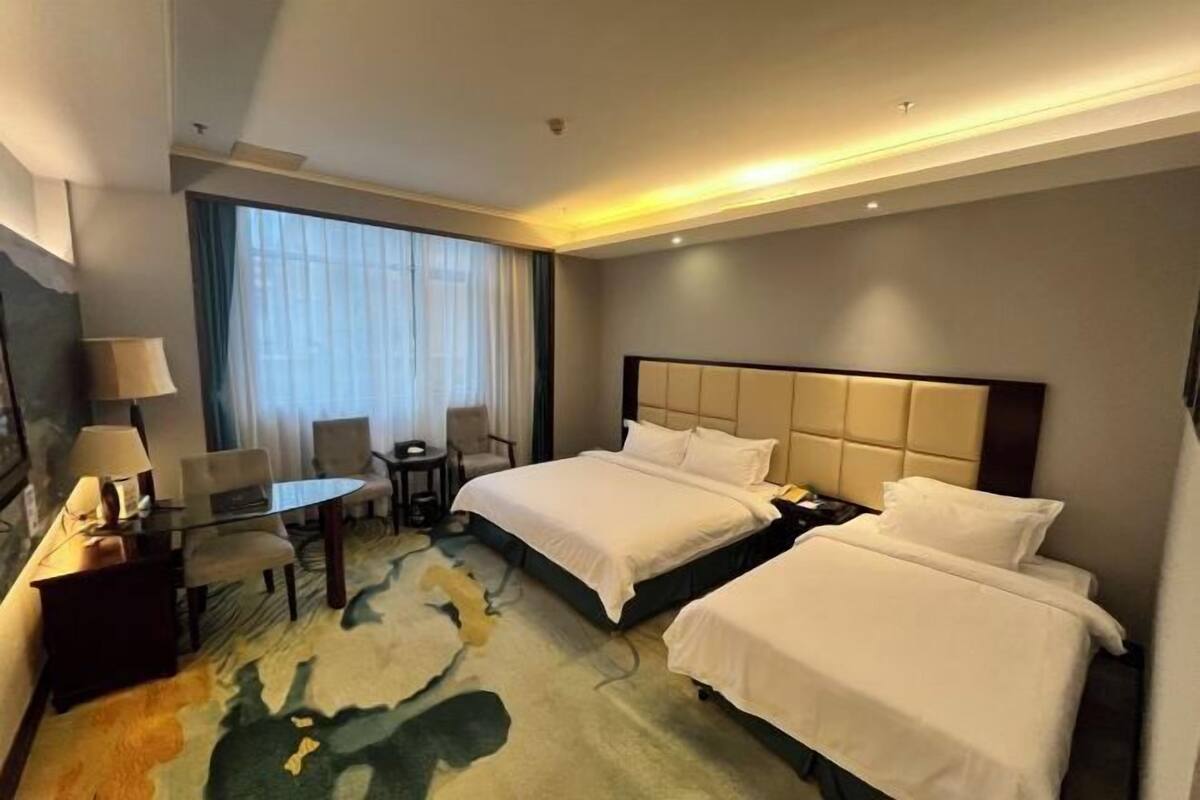 A hotel room with two beds, a desk with a chair, a small table, and a large window with curtains.