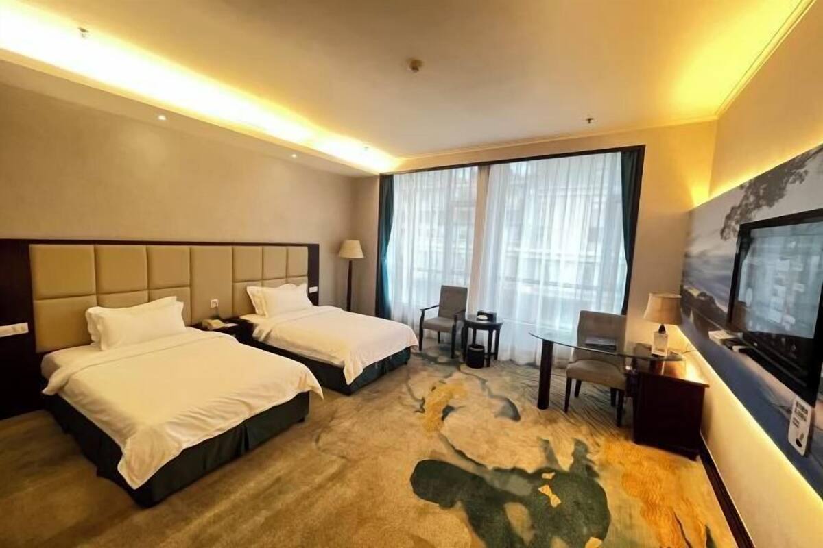 A hotel room with two beds, a desk, a chair, a television, and a large window with sheer curtains.