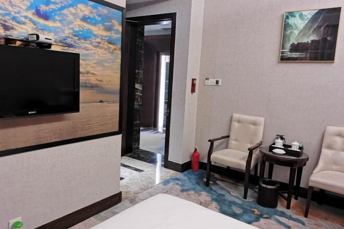 A hotel room with a TV, two chairs, a small table with items, a fire extinguisher, and a door leading to another room.