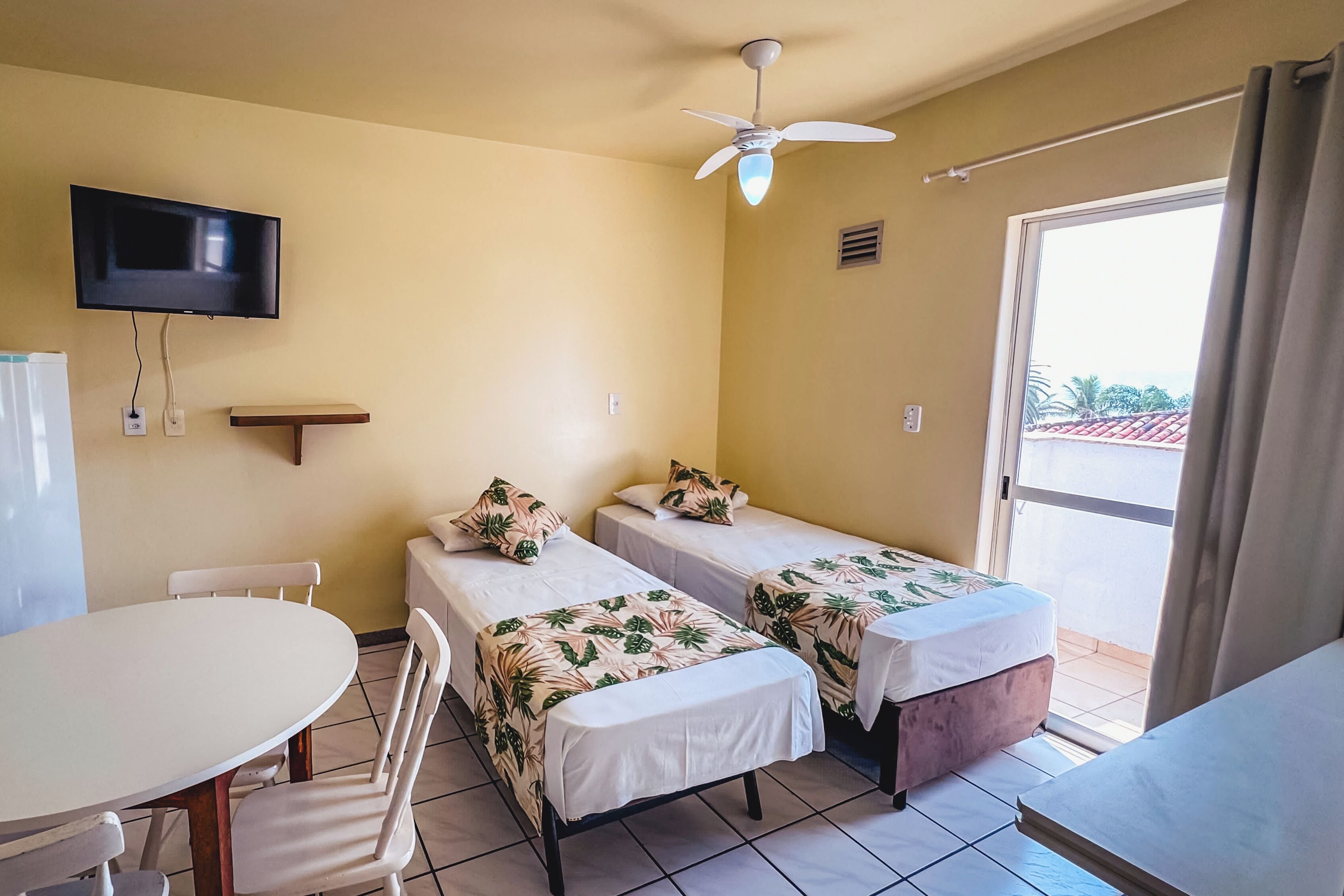 A room with two beds, a TV, a ceiling fan, a table, and a window with curtains.