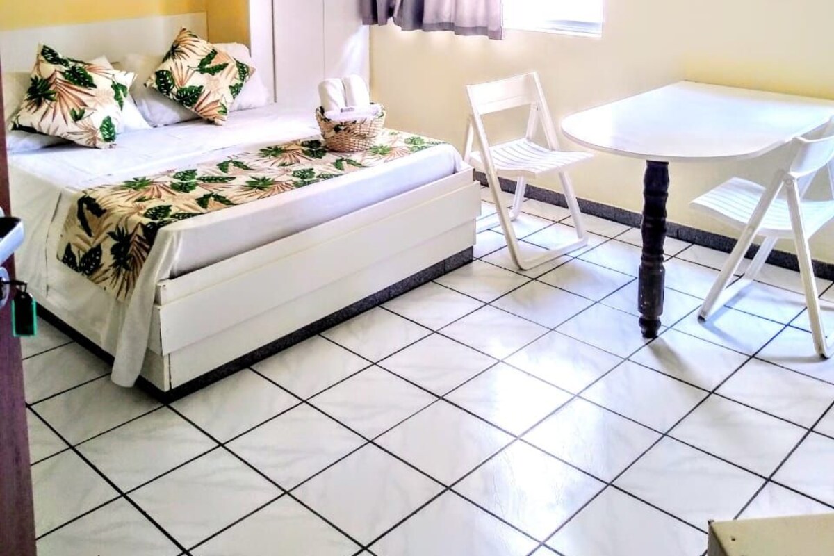 A bedroom with a bed, a small table, and a chair. There is a window with curtains, a ceiling fan, and a wall-mounted TV.